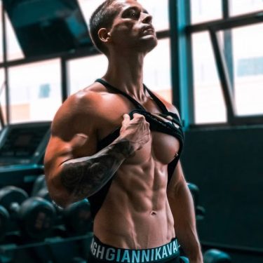 Muscular man showing abs and tattoos in a gym setting.